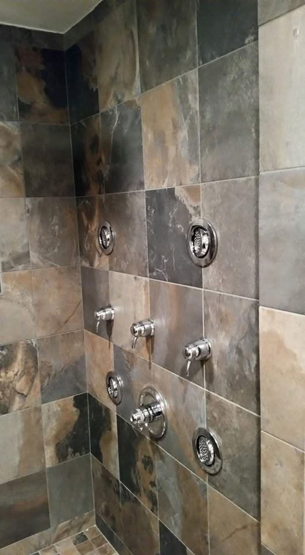 Modern shower wall with textured tiles and multiple chrome fixtures for adjustable water flow.