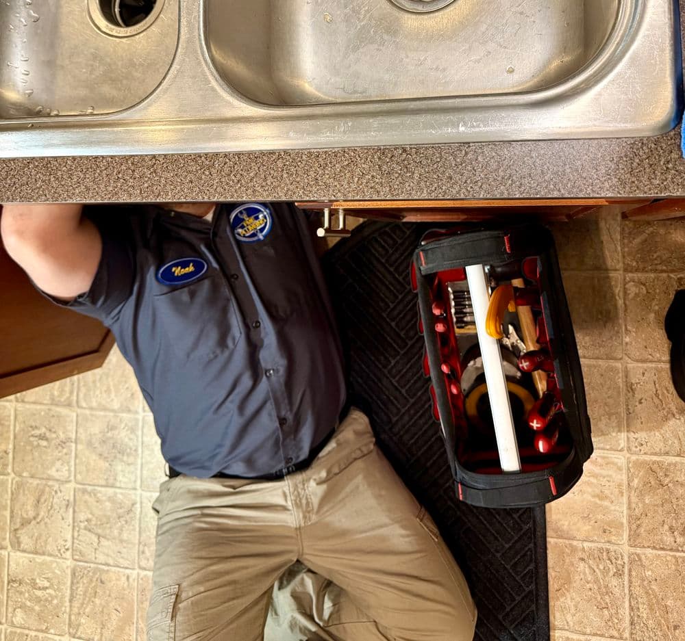 Plumber working under kitchen sink with tools in a bag and maintenance tasks in progress.