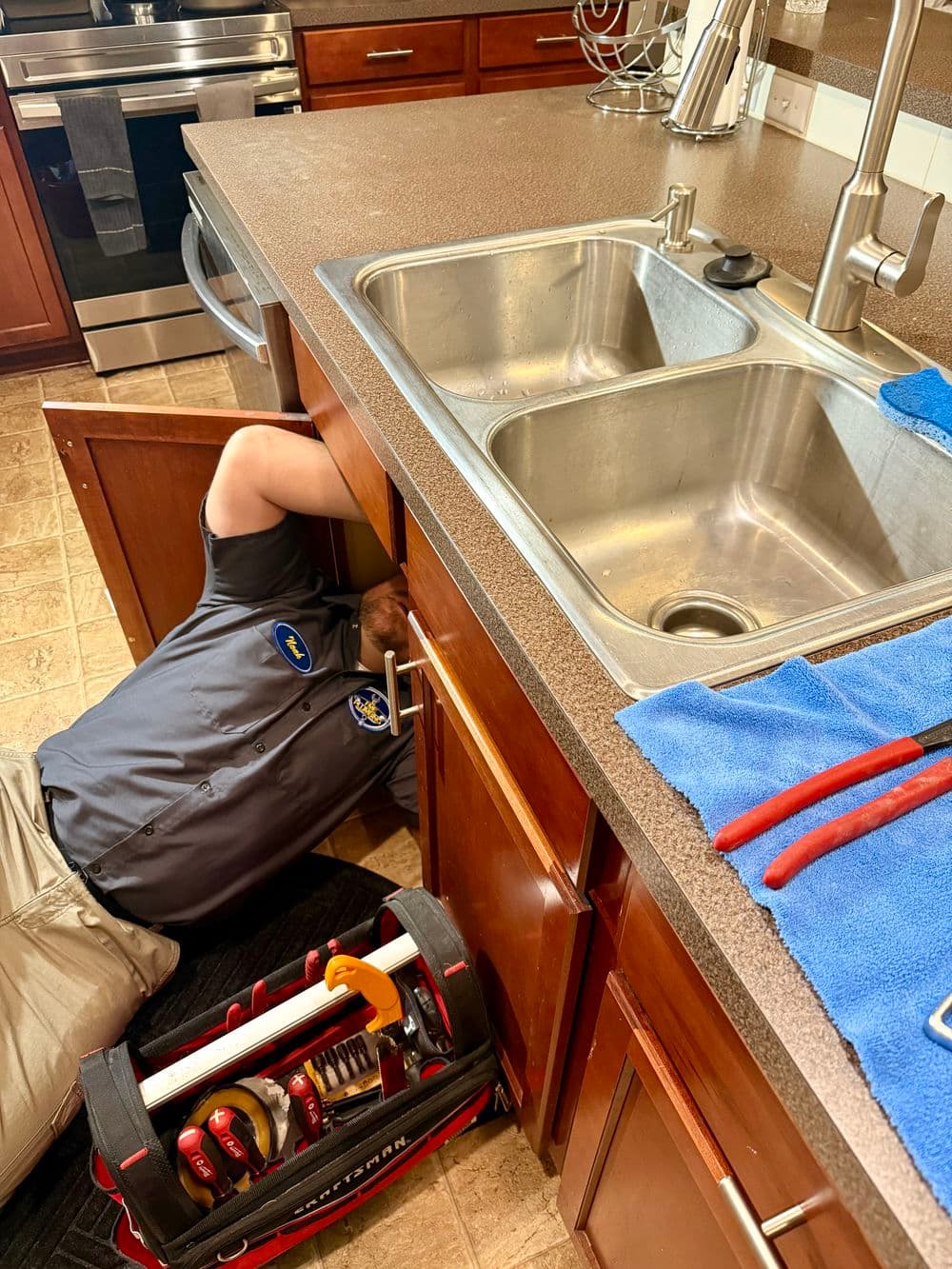 Plumber inspecting kitchen sink undercabinet with tools and repair equipment visible.